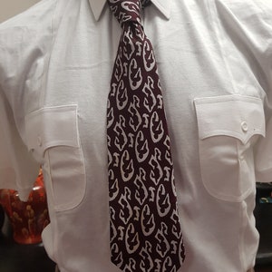 Puede incluir: Una corbata color burdeos con un patrón geométrico blanco. La corbata está anudada y se lleva con una camisa blanca de botones.