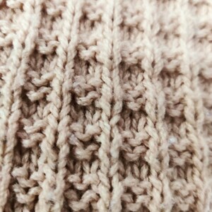May include: Close-up of a beige knitted fabric with a textured pattern. The fabric is made of soft yarn and has a warm, cozy feel.