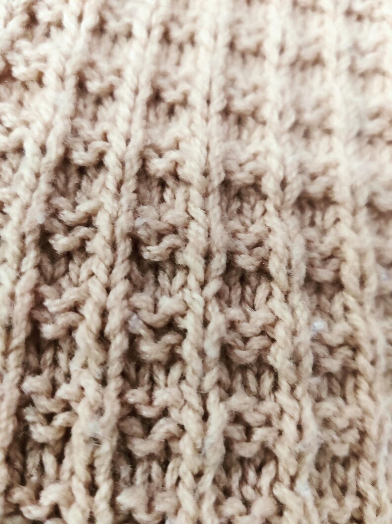 May include: Close-up of a beige knitted fabric with a textured pattern. The fabric is made of soft yarn and has a warm, cozy feel.