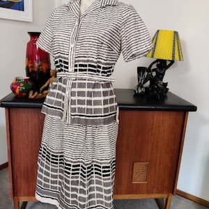 May include: A vintage dress with a white background and black and grey stripes and geometric patterns. The dress has a short sleeve, a button-up front, and a belt.
