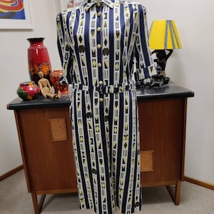 Puede incluir: Un vestido vintage con estampado de rayas azules y blancas y un diseño geométrico. El vestido tiene cuello, cierre de botones en la parte delantera y cinturón.
