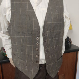 Può includere: Un gilet a quadri marroni e beige con chiusura a bottoni. Il gilet ha due tasche sul davanti ed è indossato sopra una camicia bianca con bottoni.