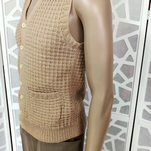 May include: A brown knitted sleeveless vest with a V-neck and button closure. The vest has a pocket on the front and a waffle-like texture.