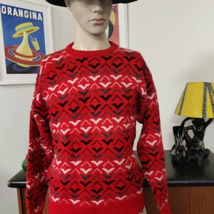 May include: Red jumper with a geometric pattern of black, white and grey diamonds and arrows. The jumper has a crew neck and long sleeves.