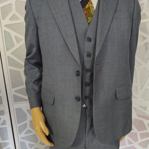 May include: A three-piece gray suit, including a jacket, vest, and trousers. The jacket has two buttons and a notched lapel. The vest has four buttons. A white dress shirt and patterned tie complete the ensemble. The suit is displayed on a mannequin.