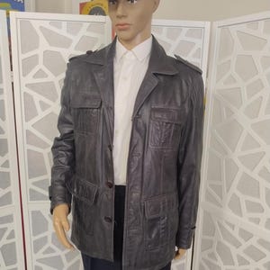May include: A dark brown leather jacket with four front pockets, button closures, and shoulder epaulets. The jacket is worn over a white collared shirt and dark pants. A brown tweed flat cap completes the outfit.