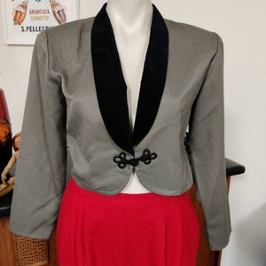 May include: A cropped grey blazer with a black velvet collar and decorative black buttons. The blazer is worn over a pair of red trousers.