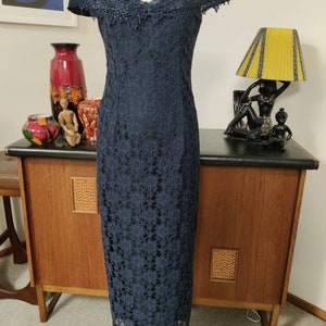 Può includere: Un abito in pizzo blu navy con scollo a balze e maniche corte. L'abito è aderente e ha una lunghezza midi.