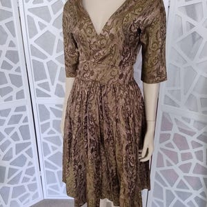 Puede incluir: Vestido de estilo vintage en tonos marrón y verde oliva, con estampado de cachemira. El vestido tiene escote en V, mangas hasta el codo y un corpiño ajustado que fluye hacia una falda acampanada. El vestido se exhibe en un maniquí.