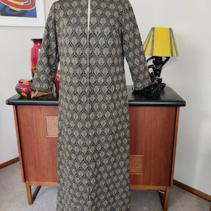 May include: A long black and gold patterned coat with a zip down the front. The coat is on a mannequin and is displayed against a wooden cabinet.