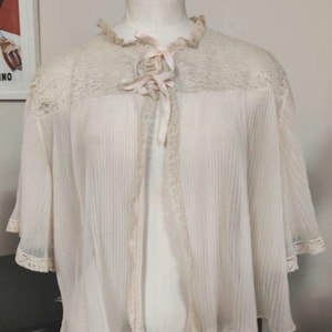 Könnte beinhalten: Ein weißes, durchsichtiges Spitzenkleid mit Rüschen am Ausschnitt und einer Schleife am Hals. Das Kleid hat ein plissiertes Design und ist aus einem leichten Stoff gefertigt.