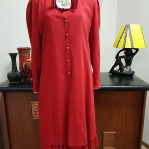 Puede incluir: Un vestido rojo con una flor blanca y un lazo rojo en el escote. El vestido tiene una falda larga con un volante en la parte inferior.