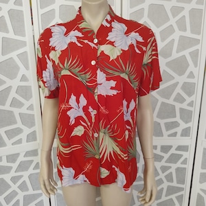 Può includere: Una camicia rossa con bottoni con un motivo floreale tropicale con foglie e fiori bianchi e verdi. La camicia a maniche corte è esposta su un manichino.