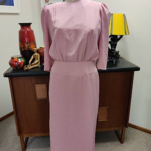 Könnte beinhalten: Ein rosa Kleid mit Rundhalsausschnitt und 3/4-Ärmeln. Das Kleid hat einen taillierten Oberteil und einen leicht ausgestellten Rock.