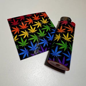 Può includere: Un accendino nero e una piccola custodia rettangolare. Entrambi gli articoli presentano un motivo ripetuto di foglie di cannabis in un arcobaleno di colori. L'accendino ha un pulsante di accensione rosso. Gli articoli sono su una superficie bianca.
