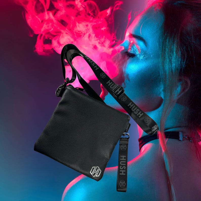 K&ouml;nnte beinhalten: Schwarze Leder-Umh&auml;ngetasche mit einem schwarzen Riemen, auf dem "Hush" in wei&szlig;en Buchstaben steht. Die Tasche hat einen silbernen Rei&szlig;verschluss und ein silbernes Logo auf der Vorderseite.