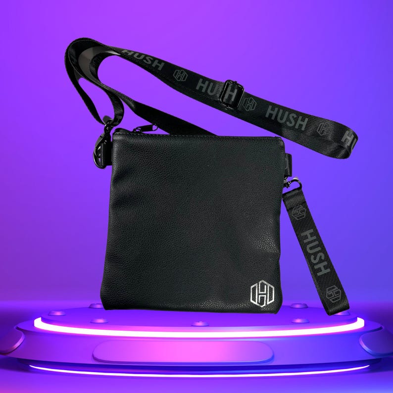 K&ouml;nnte beinhalten: Schwarze Leder-Umh&auml;ngetasche mit einem schwarzen Riemen, auf dem das Wort "HUSH" in Grau gedruckt ist. Die Tasche hat ein silbernes Logo auf der Vorderseite.