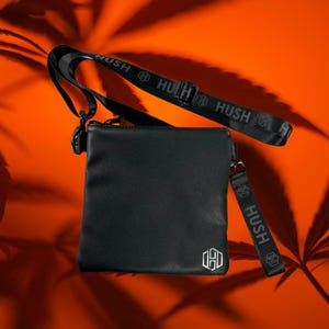K&ouml;nnte beinhalten: Schwarze Leder-Umh&auml;ngetasche mit einem schwarzen, verstellbaren Riemen, auf dem "HUSH" in wei&szlig;en Buchstaben steht. Die Tasche hat ein silbernes sechseckiges Logo mit dem Buchstaben "H" darin.