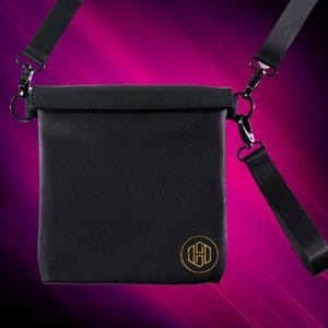 K&ouml;nnte beinhalten: Eine schwarze Leder-Umh&auml;ngetasche mit goldenem Logo. Die Tasche hat einen Klappverschluss und einen verstellbaren Riemen.