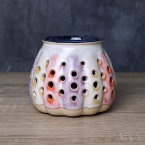 May include: A ceramic candle holder with a cream-coloured exterior, adorned with colourful perforations in orange, yellow, and purple. The top rim is navy blue. The base is a light beige colour. The candle holder is on a wooden surface.