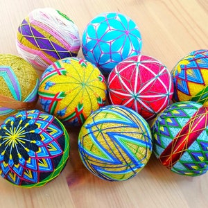 Puede incluir: Ocho bolas decorativas japonesas cosidas a mano, conocidas como 'Temari', están dispuestas sobre una superficie de madera. Las bolas presentan una variedad de diseños geométricos y florales en tonos de azul, amarillo, rojo, verde y rosa.