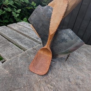 May include: Axe and wooden spoon on a tree stump. The axe handle is engraved with "Gränsfors Bruk Sweden". The spoon is light brown with a long handle. The axe head is dark grey. The items are displayed outdoors.