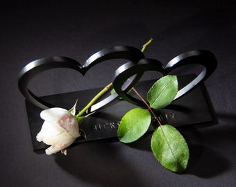 May include: Two black metal hearts intertwined with a white rose and green leaves. The base of the hearts is engraved with the names "Nick &" and the year "2017".