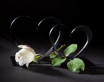 May include: A black metal sculpture of two intertwined hearts, with a white rose and green leaves resting on a dark surface. The sculpture is a symbol of love and romance, perfect for a wedding or anniversary gift.