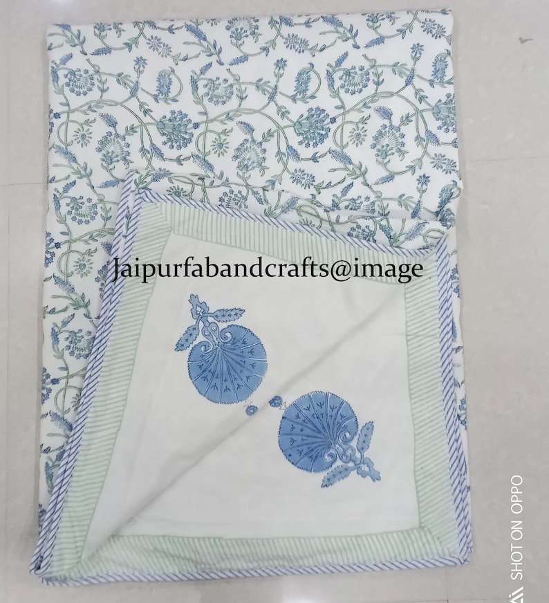 May include: A white cotton quilt with a blue and green floral print. The quilt has a light green border with white stripes. The quilt is folded in half and the inside of the quilt has a blue and white floral print.