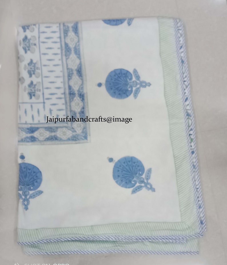May include: White cotton fabric with a blue and white floral print. The fabric has a green border with a white stripe. The text "jaipurfabandcrafts@image" is printed on the fabric.