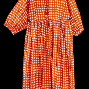 Può includere: Abito a quadri arancione e bianco con maniche a tre quarti. L'abito ha uno scollo tondo con fiocco e vita arricciata. È appeso a una gruccia blu con motivo floreale.