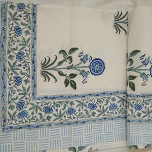Puede incluir: Tela de algodón blanca con estampado floral azul y verde. La tela tiene un borde con un patrón repetitivo de rayas azules y blancas.