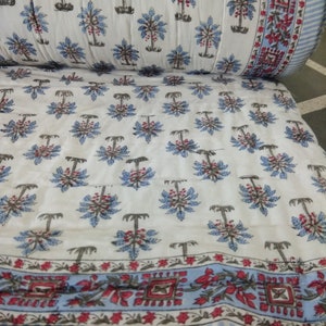 Op de afbeelding: Witte katoenen stof met een blauwe en rode bloemenprint van palmbomen. De stof heeft een blauw-witte gestreepte rand met een rood-wit bloemenontwerp.
