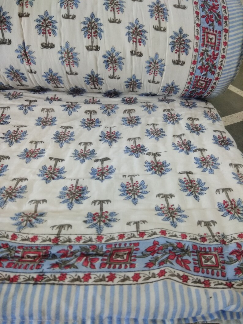 Op de afbeelding: Witte katoenen stof met een blauwe en rode bloemenprint van palmbomen. De stof heeft een blauw-witte gestreepte rand met een rood-wit bloemenontwerp.