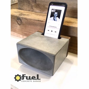 May include: A gray concrete phone stand with a wood accent. The stand has a curved opening to amplify sound. The stand is holding a smartphone playing music. The phone screen displays the song title "A Certain Romance" by The Smiths.
