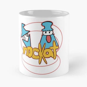 Könnte beinhalten: Eine weiße Keramiktasse mit einer Cartoon-Illustration von zwei blauen Figuren und dem Wort "nockat" in gelben Buchstaben.