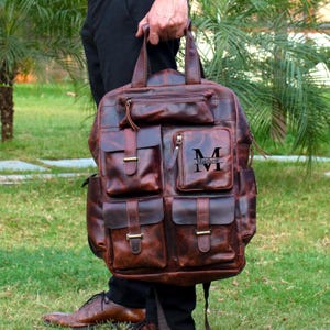 May include: A brown leather backpack with multiple pockets and compartments. The backpack has a top handle and shoulder straps. The front of the bag features the initials "M" and "IVI" with the name "Matthew" below. The bag is held by a person.