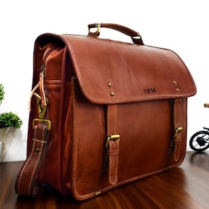 May include: A brown leather satchel with a top handle, shoulder strap, and buckle closures. The bag features the initials "SPM" on the front flap. The bag is approximately 14 inches wide and 11 inches tall.