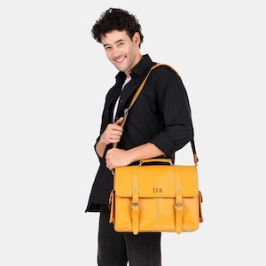 May include: A mustard yellow leather messenger bag featuring a top handle, an adjustable shoulder strap, two front buckle closures, and side pockets. The front flap is embossed with the black capital letters IJA in a serif font.