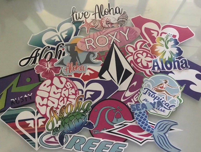 May include: A collection of colorful stickers featuring various designs, including a turtle, a mermaid, a hibiscus flower, and the words "Live Aloha", "Roxy", "Sea Ya", "Tropical Paradise", and "Rusty".