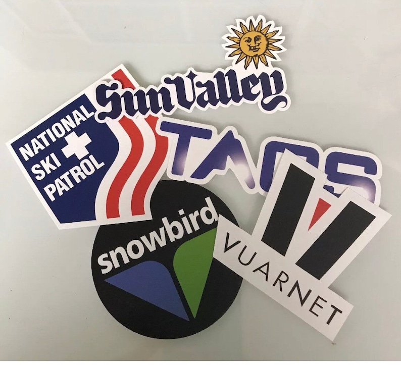 May include: A collection of colorful ski resort stickers, including Sun Valley, Taos, Snowbird, and Vuarnnet. The stickers feature a variety of designs, including a sun with a face, a mountain range, and a ski patrol logo.
