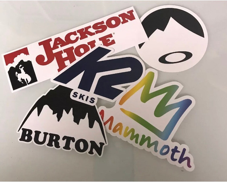 May include: Assortment of colorful stickers featuring various ski resort and outdoor brand logos, including Jackson Hole, K2 Skis, Burton, Mammoth Mountain, and a logo with a mountain silhouette and a circle.