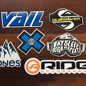 Puede incluir: Una colecci&oacute;n de siete pegatinas de snowboard de colores. Las pegatinas presentan logotipos de marcas como Vail, Quiksilver, Jones, Ride Snowboards, y una pegatina con el texto "Eat Sleep Ride Repeat" en unas gafas.
