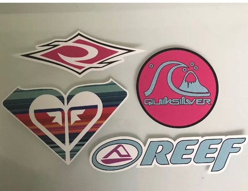 May include: Four colorful stickers with surf brand logos. The stickers are on a white background. The logos are Quiksilver, Reef, and two different variations of the Roxy logo.