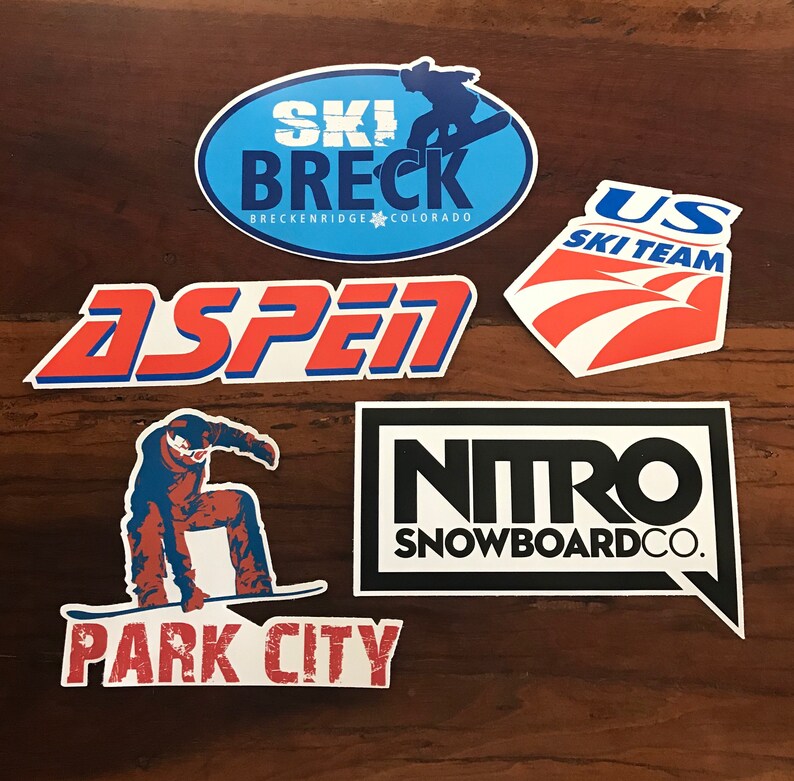 Puede incluir: Cinco pegatinas coloridas con logotipos de estaciones de esqu&iacute;. Las pegatinas est&aacute;n sobre una superficie de madera. Los logotipos son "Ski Breckenridge Colorado", "Aspen", "US Ski Team", "Nitro Snowboard Co." y "Park City".