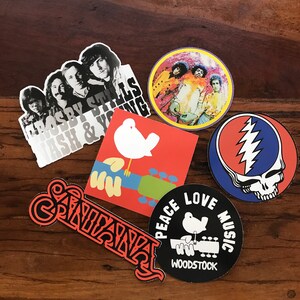 May include: A collection of eight vintage music-themed stickers. The stickers feature various musicians, bands, and music festivals, including Crosby, Stills & Nash, The Grateful Dead, and Woodstock. The stickers are printed on a white background with colourful designs.