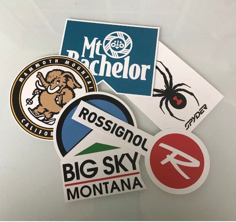 May include: Five colorful stickers with different designs. The stickers feature a black spider with red accents, a blue and white mountain logo with the text "Mt Bachelor", a brown elephant with the text "Mammoth Mountain California", a blue and green mountain logo with the text "Rossignol Big Sky Montana", and a red circle with a white "R" inside.