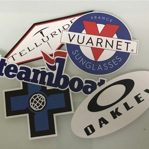 May include: Five different stickers with various logos. The stickers are white with black, blue, red, and white lettering. The logos include "Telluride", "Steamboat", "Oakley", and "Vuarnent Sunglasses, France".
