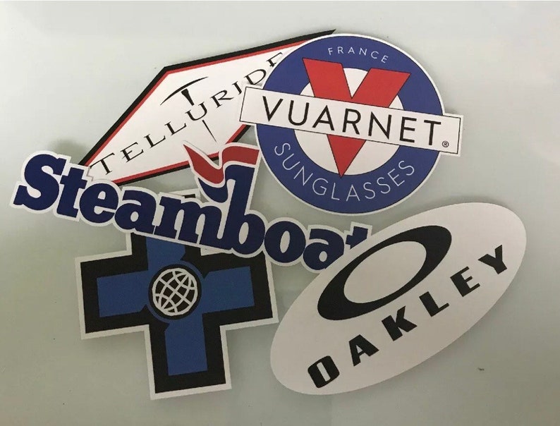May include: Five different stickers with various logos. The stickers are white with black, blue, red, and white lettering. The logos include "Telluride", "Steamboat", "Oakley", and "Vuarnent Sunglasses, France".