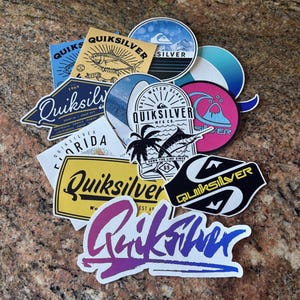 Puede incluir: Una colección de 11 pegatinas de la marca Quiksilver con varios diseños, que incluyen un pez, una palmera, una ola y el logotipo de Quiksilver. Las pegatinas están sobre una superficie marrón texturizada.